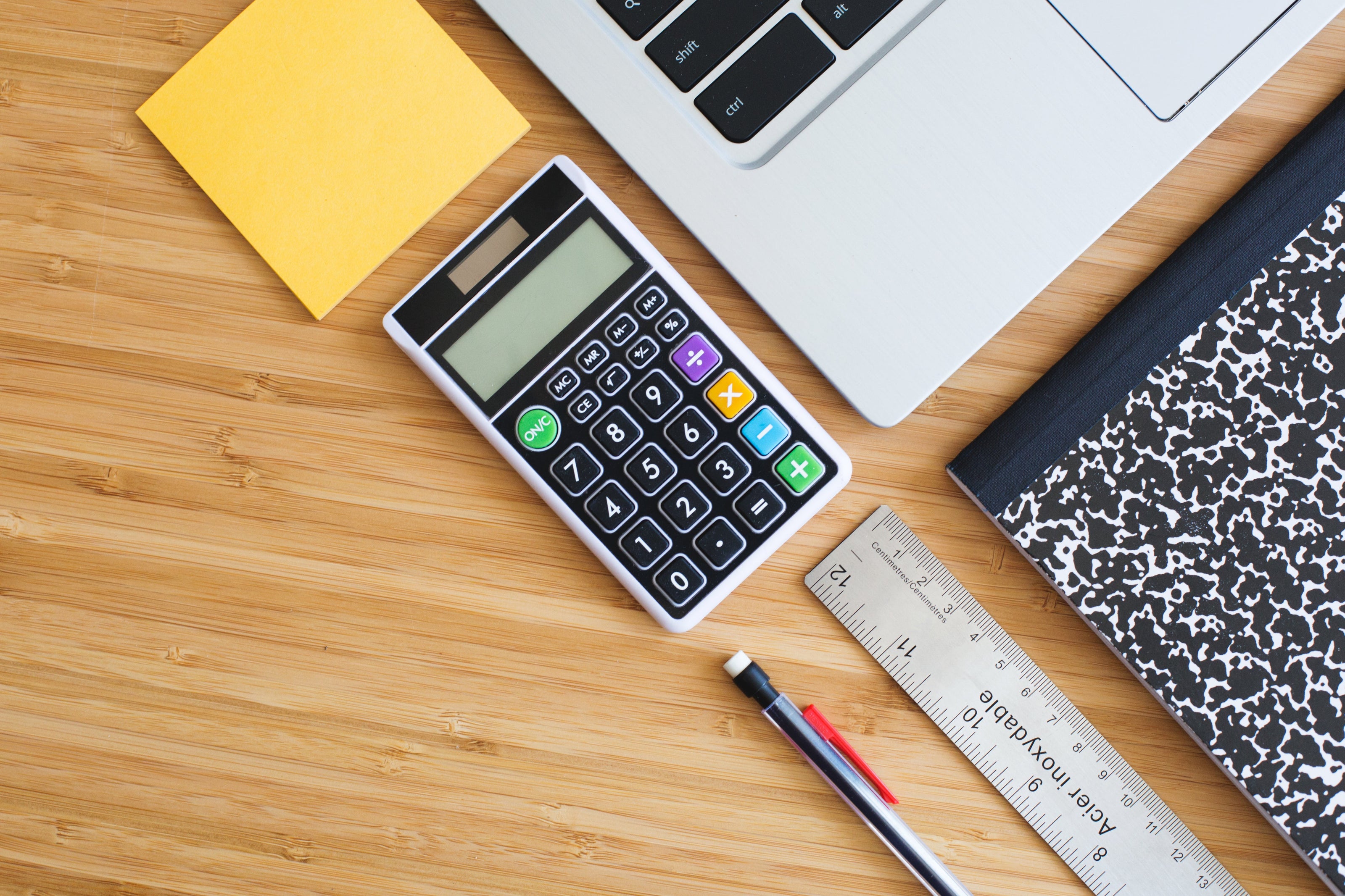 A professional woman writes on a notepad in an office, keeping organised in work. Image focuses on a clean desk, a stationary organiser tumbler with pencils, a glass of water, a smart phone, a notepad, and a professional's hands writing in calligraphy-style. 